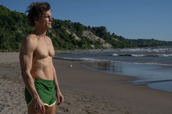 Hawk (Matt Bomer), in green short shorts, stands on a beach